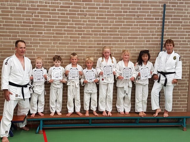 Zeven jonge judoka’s in witte judopakken staan trots op een bank in de gymzaal, elk met hun examendiploma in de hand. Links en rechts staan hun judoleraren met een zwarte band.